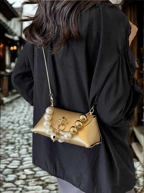 Person wearing a black coat with a gold handbag featuring pearl embellishments on a cobblestone street.
