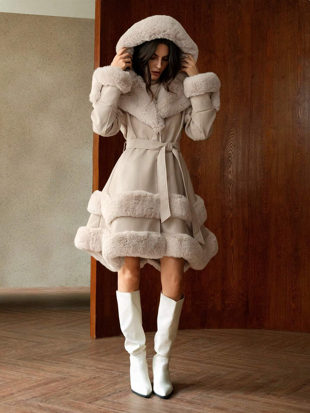 Woman wearing a beige coat with fur trim and white boots in a room with wooden flooring and a textured wall.